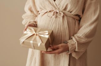 Gestante com vestido bege segurando caixa de presente com laço claro nas mãos