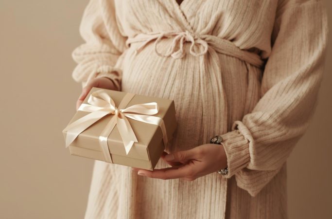 Gestante com vestido bege segurando caixa de presente com laço claro nas mãos
