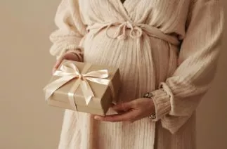 Gestante com vestido bege segurando caixa de presente com laço claro nas mãos