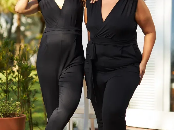Duas mulheres usando macacão para gestante preto com decote transpassado, modelagem confortável e faixa na cintura, em look casual com sandália e tênis.